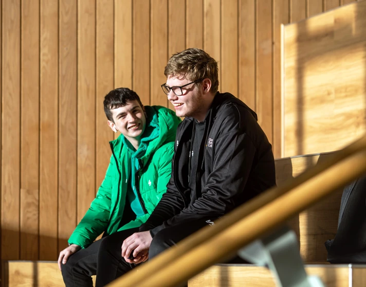 Two students sitting on tiered wooden seating at Springburn campus, chatting and enjoying the sunlight. Two students sitting on tiered wooden seating at Springburn campus, chatting and enjoying the sunlight.